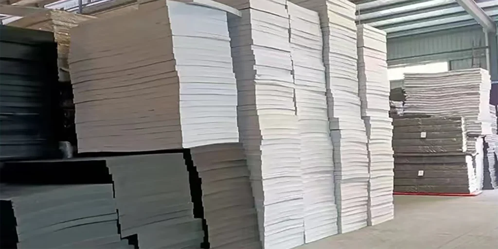 a large warehouse interior showcasing substantial stacks of foam sheets. The sheets are primarily in white and gray tones, neatly piled in tall, orderly stacks. The warehouse features a spacious, industrial - style setting with a metal roof structure visible overhead. These foam materials are likely used for applications such as product packaging, cushioning, or thermal insulation in various industrial and manufacturing processes.