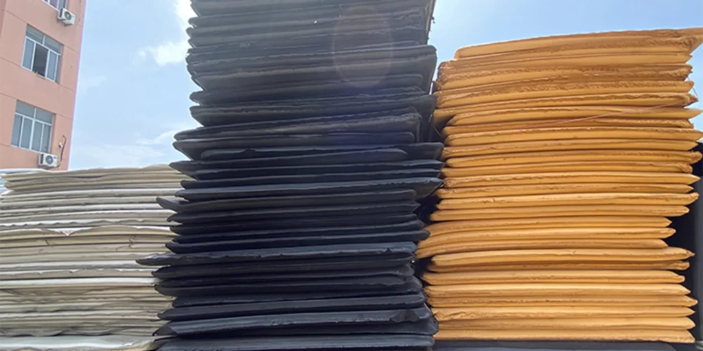 a large number of stacked materials, which appear to be sheets or panels. There are three distinct colors visible: white on the left, black in the middle, and brown on the right. The materials have a uniform thickness and seem to be made from some type of flexible material, possibly rubber or plastic. They are neatly arranged in vertical columns, with each column having multiple layers. In the background, there is a building with windows and an air conditioning unit, suggesting that this might be an outdoor storage area near a commercial or industrial facility.