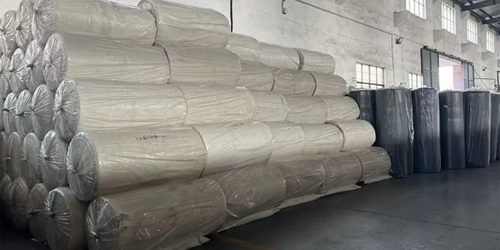 a warehouse or storage facility with large rolls of material stacked in an orderly fashion. These rolls are wrapped in transparent plastic, likely for protection and ease of handling. The materials appear to be textiles or fabrics, given their soft appearance and the way they are rolled up.On the left side of the image, there is a prominent stack of these rolls, which seems to be the main focus of the photo. To the right, additional rolls can be seen, though not as closely packed or as tall as the central stack.The background features industrial elements such as metal beams and windows that allow natural light into the space. There’s also what appears to be a door at the far end of the room, suggesting access points for loading and unloading goods.Overall, the scene conveys a sense of orderliness and efficiency typical of a well-maintained storage area within a larger manufacturing or distribution center.