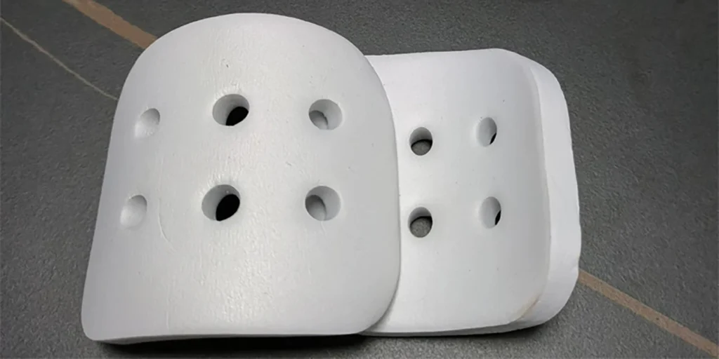 two white protective pads placed on a gray surface. Each pad features multiple circular holes, which may help with breathability and reduce weight. Their smooth, rounded shape suggests they could be used as shin guards, knee pads, or other protective gear in sports like soccer, hockey, or martial arts. The minimalist design focuses on functionality, ensuring protection while allowing airflow.