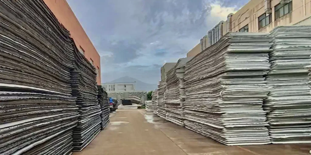 a vast industrial or commercial storage area, characterized by towering stacks of flat, rectangular objects. These items are meticulously arranged in neat rows and columns, extending from the foreground to the background of the scene.On either side of the central pathway, which is flanked by these orderly piles, there are buildings with neutral-colored facades that suggest an operational facility possibly related to manufacturing, warehousing, or distribution. The sky above is overcast, hinting at cloudy weather conditions, yet no rain appears to be falling as the ground does not show signs of wetness.In the distance, beyond the immediate focus on the stacked materials, one can observe additional structures and what seems like a mountain range under the heavy clouds, adding depth to the landscape captured in this photograph.