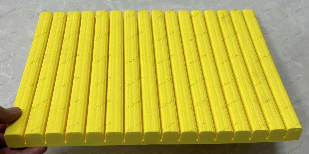 a collection of uniformly shaped, bright yellow objects arranged in rows on what appears to be a white surface. Each object has a cylindrical shape with ridges running along its length, giving it a textured appearance. The ends of the objects are rounded and appear smooth compared to the rest of their bodies. A hand is visible at the bottom left corner of the image, providing a sense of scale for the size of these objects. They seem to be made from a material that reflects light softly, suggesting they could be plastic or a similar synthetic substance. The precise purpose of these objects isn’t immediately clear without additional context, but given their uniformity and construction, they might serve as components in some sort of assembly or perhaps as parts for a specific tool or device. Their vibrant color also suggests they may have been chosen for visibility purposes, possibly for safety reasons if used in an industrial setting.