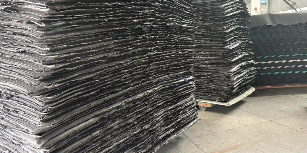 an indoor warehouse setting where massive stacks of black industrial sheets are stored. The sheets are layered in large piles, and the warehouse features a concrete floor. In the background, there are industrial structures, including a cart with more stacks of similar sheets and other industrial materials. The scene indicates a storage or manufacturing environment for these large, black industrial components.