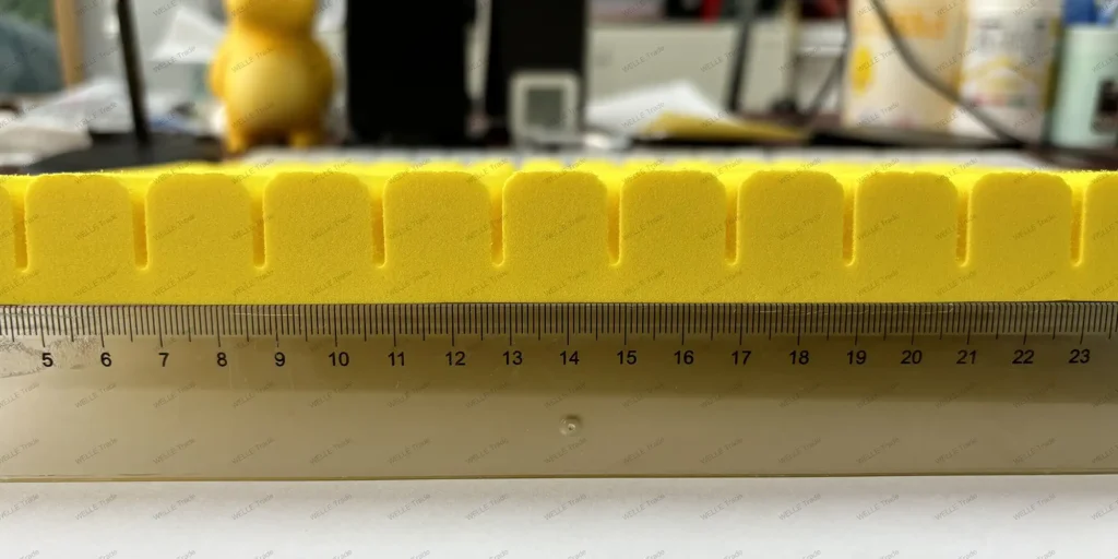 a close-up view of an array of yellow, foam-like objects neatly aligned on a flat surface. Each object has a distinct shape with a rounded top and a rectangular base, creating a visually appealing pattern. The precise arrangement suggests that these items are part of a larger set or collection. In the foreground, there is a transparent ruler placed horizontally beneath the row of yellow objects, providing a scale for measurement. The ruler’s markings indicate centimeters, which helps to gauge the size of each item in the series. The background features a blurred assortment of various items, including what appears to be office supplies, containers, and possibly some electronic devices. This setting gives the impression that the photo was taken in a workspace or laboratory environment where precision and organization are key elements.
