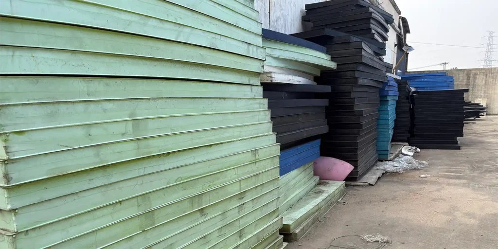 an outdoor industrial storage area where multiple tall stacks of large plates in different colors, including green, black, and blue, are arranged. The plates appear to be made of a sturdy material, likely for industrial or manufacturing purposes. The ground is concrete, and there are some scattered items and industrial structures visible in the background, indicating a functional storage or production environment.