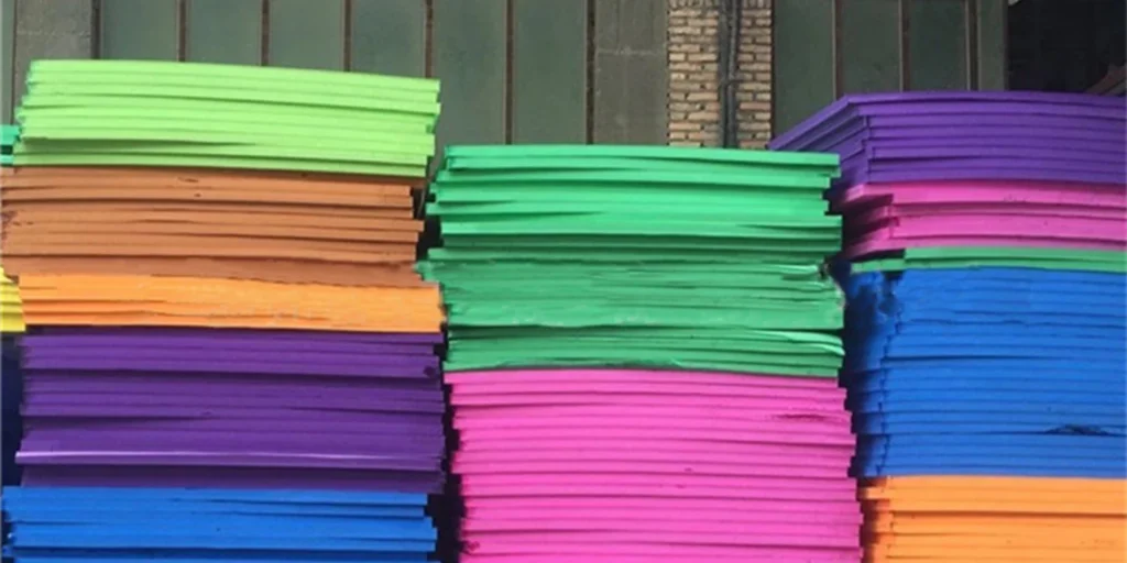 a collection of colorful, rectangular objects stacked in rows and columns. The colors include shades of green, purple, blue, orange, pink, and brown. Each stack appears to be made up of multiple layers, creating a vibrant mosaic effect. The background is nondescript with a neutral color palette that contrasts with the bright hues of the foreground items.