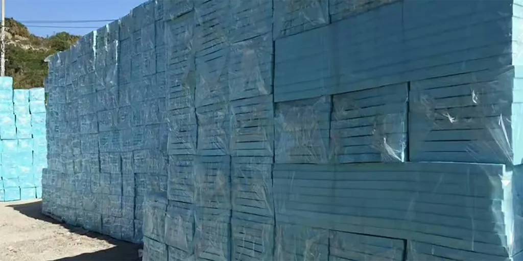 a substantial quantity of blue - colored insulation boards, neatly stacked in an outdoor setting. Each board is wrapped in clear plastic, likely for protection during storage and transportation. The stacks are tall and arranged in rows, occupying a gravel - covered ground area. In the background, there are more stacks of the same blue insulation boards, and a distant hill with some vegetation under a clear blue sky, suggesting an open - air storage yard, probably associated with a construction materials supply business. These insulation panels are commonly used in building projects for thermal insulation in walls, roofs, or floors.