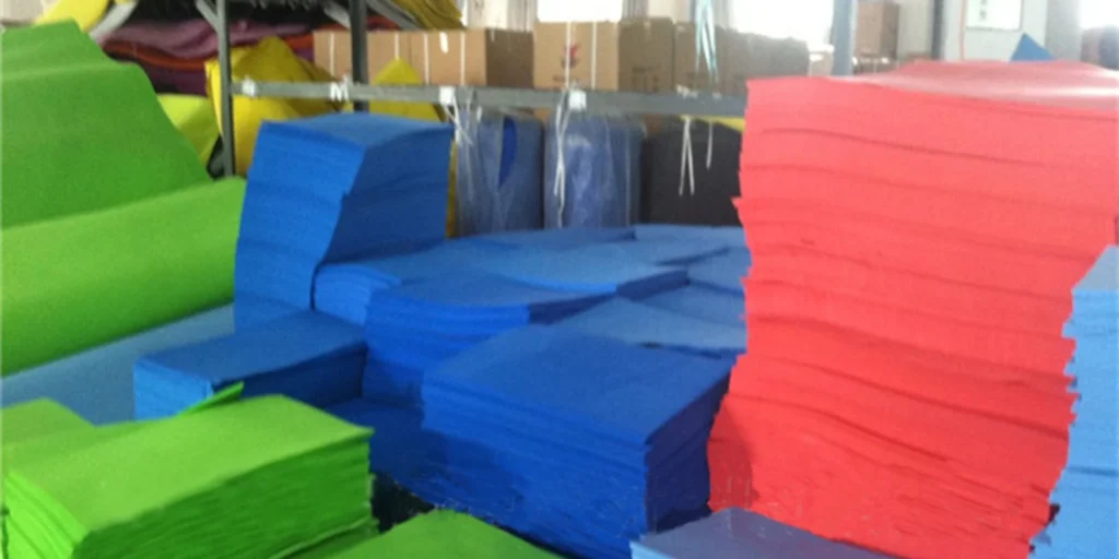 a warehouse or storage area filled with various colored foam mats. These mats are stacked in different arrangements, showcasing an array of vibrant colors including red, blue, green, and purple. The mats appear to be made from a soft, flexible material suitable for use as exercise mats or protective flooring.In the background, there is shelving that seems to hold additional items, possibly more mats or other types of materials. The lighting suggests it could be daytime, given the brightness and shadows cast by the light source. Overall, the scene gives off an industrial feel, typical of a space used for storing goods before distribution or sale.