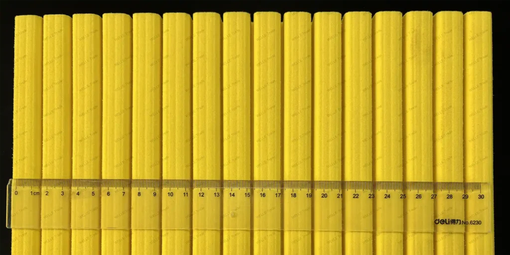 a collection of uniformly shaped, yellow objects arranged in neat rows. Each object has a cylindrical form with ridges running along its length, giving it a textured appearance. The consistent color and design suggest that these items are likely part of a set or produced for a specific purpose. In the foreground, there is a transparent ruler placed horizontally across the middle row of the objects. This ruler provides a scale to measure the size of the objects, which appear to be approximately 30 centimeters long based on the visible markings from 0 to 30 centimeters on the ruler. The ruler itself seems to have fine gradations, allowing for precise measurements. The background is dark, providing a stark contrast to the bright yellow hue of the objects, making them stand out prominently. There’s also some text printed on the bottom right corner of the ruler, but due to the resolution constraints, the details cannot be discerned clearly.