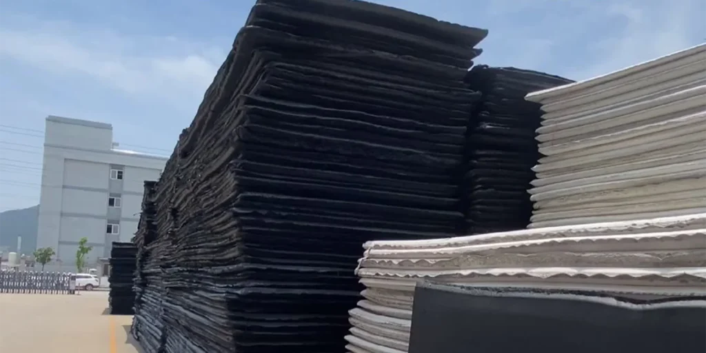 a large number of stacked materials, which appear to be sheets or panels. On the left side, there are tall stacks of black material, while on the right side, there are shorter stacks of white material. The background features an industrial setting with buildings and a clear sky. There is also a vehicle parked in front of the building.