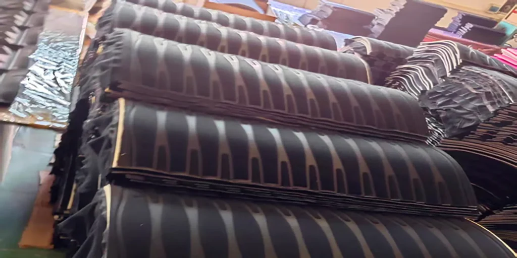multiple stacks of patterned materials in what appears to be a factory or warehouse. The materials are predominantly black with gray, striped patterns. They are neatly piled, suggesting they are ready for production or distribution. The background includes other industrial items, indicating a manufacturing environment. These materials could be used for various products, like protective gear or components in larger goods.