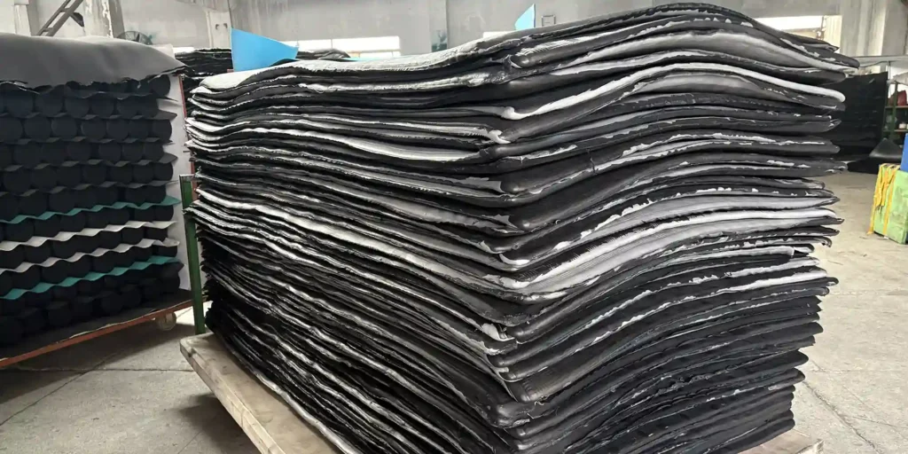 an indoor warehouse scene where a massive stack of black industrial sheets is stored on a wooden pallet. The sheets are layered in large, wavy piles. The warehouse features a concrete floor, and in the background, there are other industrial materials, including a rack with black cylindrical items and some colorful equipment. The scene indicates a storage or manufacturing environment for these large, black industrial components.