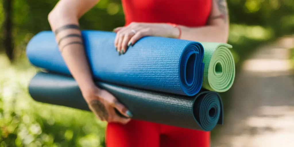 Yoga mats, female in red tights and green pants standing on grass with three yoga mats in her hands. She is wearing black tights and holding yoga mats. The background is a forest or park. The yoga mats are floating in the grass, surrounded by trees. The female is holding yoga mats and appears to be practicing yoga.