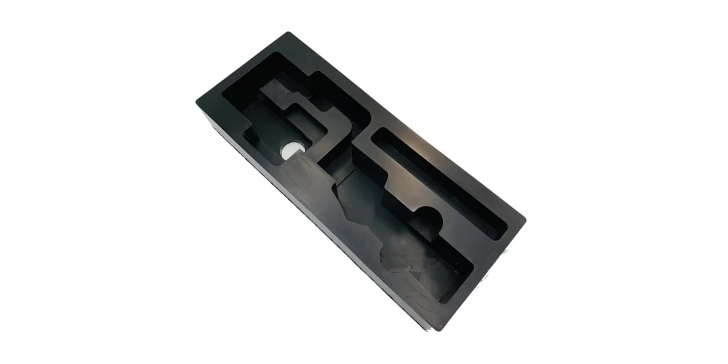 shows a long, rectangular black foam insert with a series of precision-machined cavities. The design includes a complex, shaped compartment on the left, a narrow slotted section on the right, and a recessed lower area, all tailored to fit specific items securely. The smooth, matte black finish and custom-fit layout make it ideal for organizing and protecting tools or components during transport or storage.