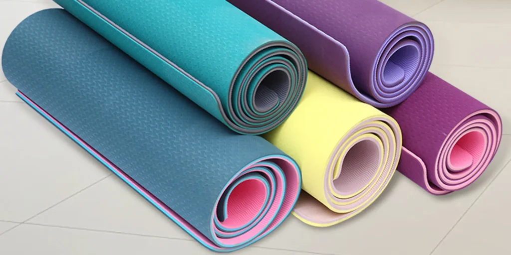 five different colors of yoga mats, they are: dark blue, light blue, purple, yellow and pink. These five yoga mats are neatly arranged on a white floor tile. They are rolled up and placed side by side. The color of the inside of the yoga mat is lighter than the outside.