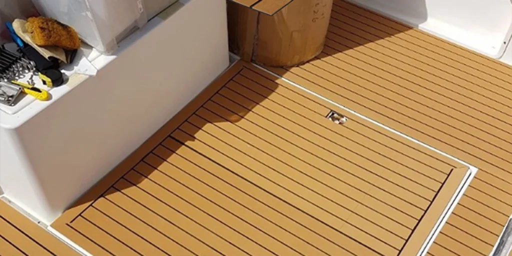 a close-up view of the deck area on what appears to be a boat or yacht. The deck is made up of wooden planks arranged in parallel lines, creating a textured and durable surface suitable for marine environments. On the left side of the image, there’s a white fixture with various items placed inside it. These include what seems to be a yellow sponge, some metal objects that could be tools or hardware, and possibly other small items not clearly visible due to the angle and resolution of the photo. In the center-right part of the image, we can see a cylindrical object which looks like a hatch cover, likely providing access to storage space or equipment below the deck level. It has a handle mechanism for opening and closing. Overall, the scene suggests maintenance work or preparation for an activity involving the use of these tools and materials.