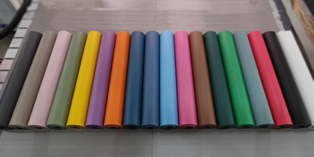 a collection of colorful rolls of paper neatly arranged in rows on a shelf. Each roll has a different color, including shades like grey, pink, yellow, orange, blue, light blue, brown, dark green, green, grey-green, red, black, and white. These papers are likely used for various purposes such as gift wrapping, crafting, or decoration. The background appears to be a store or warehouse setting with a tiled floor.