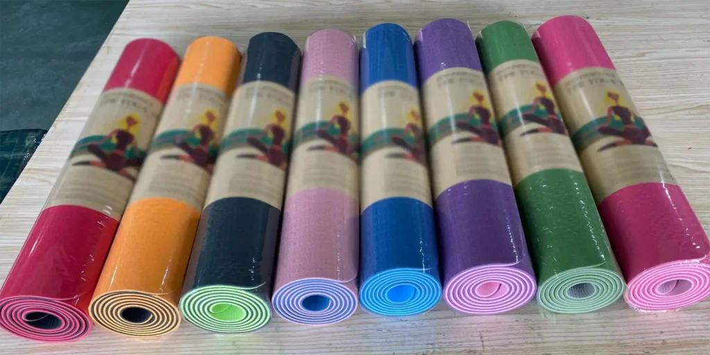 nine yoga mats rolled up and placed side by side on a wooden surface. From left to right, they come in a variety of vibrant colors: red, orange, black, pink, blue, purple, green, and two shades of pink. Each mat is wrapped in plastic packaging, which features a label with a woman practicing yoga. The labels also include some text information, but it's not clearly legible from this angle. The overall impression is neat and orderly, showcasing a range of options for potential buyers.