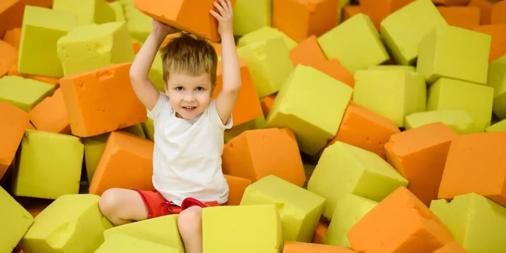 The picture shows a young boy, wearing red pants and with his hands above his head, playing in a recreational area filled with yellow and orange foam blocks.