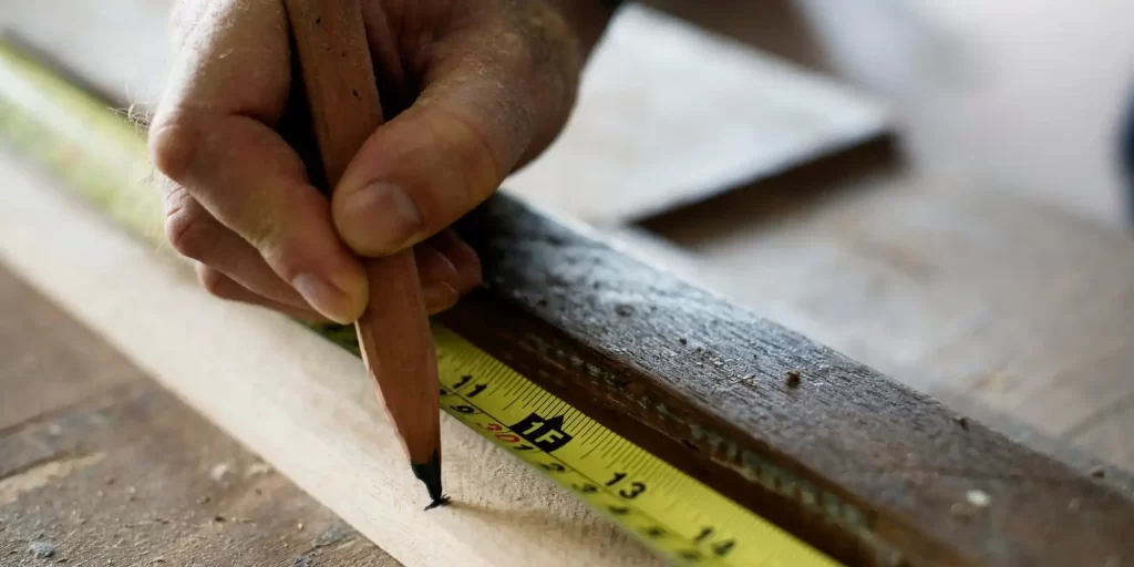a person marking a measurement on a piece of wood. A ruler is laid across the wood, and the individual is using a pencil to make the mark, indicating precision and attention to detail in woodworking or carpentry. The main focus is on a hand holding a pencil, marking on a piece of wood. A yellow measuring tape with black numbers and markings is visible, indicating precise measurements. The person’s hand appears experienced or aged, suggesting skill or expertise. The surface of the wood looks rough and unfinished. A metal ruler or straight edge tool rests on the wood parallel to the measuring tape aiding in making an accurate mark.