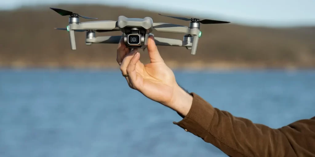 A drone is held in the hand of a man who is wearing a brown jacket as the drone hovers in the air.