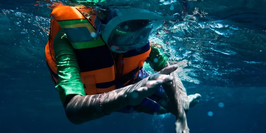 a person engaged in underwater activities, possibly snorkeling or shallow diving. The individual is wearing a bright orange life jacket, which is an essential safety device that provides buoyancy and helps the wearer stay afloat. The life jacket is likely to contain foam for buoyancy—potentially EVA foam, which is characterized by its durability, resistance to water, and excellent flotation properties.