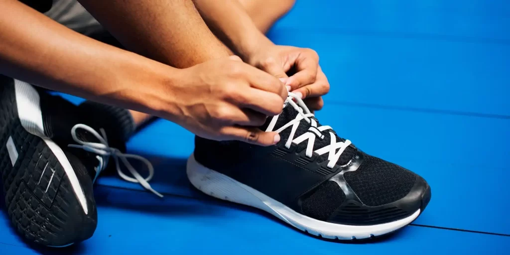 a close-up of a person tying the laces of a black sports shoe with white soles, which appears to be designed for athletic activities, possibly a type of sneaker or cleat given the context. The individual is seated on a bright blue surface, which could be indicative of a gym or sports environment. The focus on the hands tying the shoe draws attention to the act of preparation before engaging in a sport or physical activity
