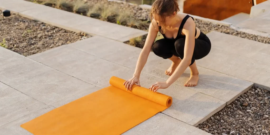 a person in workout attire rolling out an orange yoga mat on a paved outdoor surface. This scenario typically represents the setup for an outdoor exercise session, possibly yoga or a stretching routine. The title, "EVA Foam Enhancing Safety and Comfort in Mats and Walkways," suggests that the yoga mat is likely made of EVA foam, a material known for its cushioning properties, providing safety and comfort during physical activities. EVA foam is durable, slip-resistant, and often used in sports equipment for its shock-absorbing qualities.