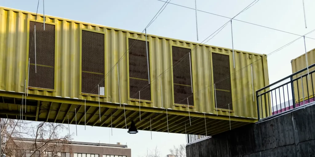 a suspended structure that appears to be a pedestrian bridge or walkway constructed from modified shipping containers. The containers are yellow, with several areas cut out and covered with what seems to be metal mesh or grating, possibly for ventilation or to serve as windows. The walkway is supported by cables or rods, suggesting a modular design that allows for relatively quick assembly. This kind of structure could be part of a temporary installation or a creative architectural solution for pedestrian traffic. The containers’ robust build makes them suitable for repurposing into functional spaces like offices, cafes, or, as shown here, walkways.