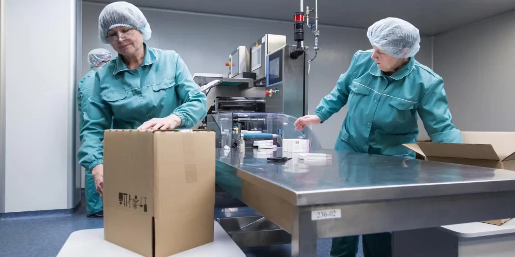 The picture shows two women working in blue uniforms, they are wearing hats and masks. On the table is a cardboard box containing products.
