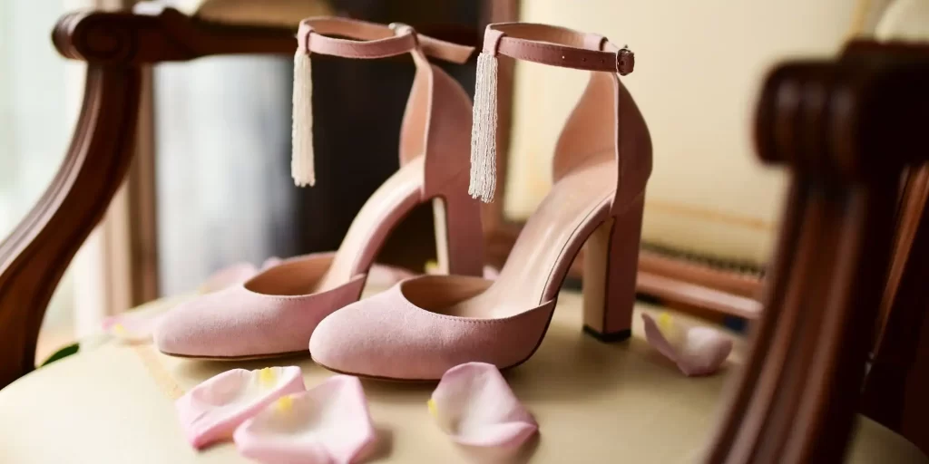 a pair of pink high-heeled shoes with tassels on the ankle straps, displayed on a chair. The shoes are surrounded by rose petals, which adds a decorative, romantic element to the scene. It appears to be a setting that could be part of a wedding preparation or a sophisticated event.