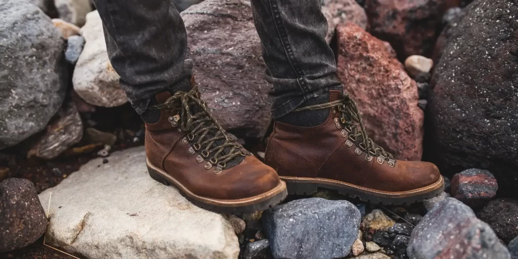 One foot is wearing a pair of brown leather boots and the other foot is next to the boots. The background is rocks and stones.