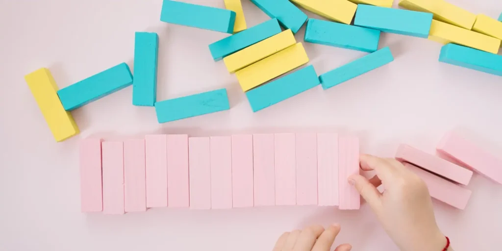 Some brightly colored blocks, mainly pink and blue. These blocks are placed on a light colored background. In the picture, a hand is picking up a blue colored block. The blocks appear to have a hard texture and a smooth surface. The overall look gives a clean and crisp look.