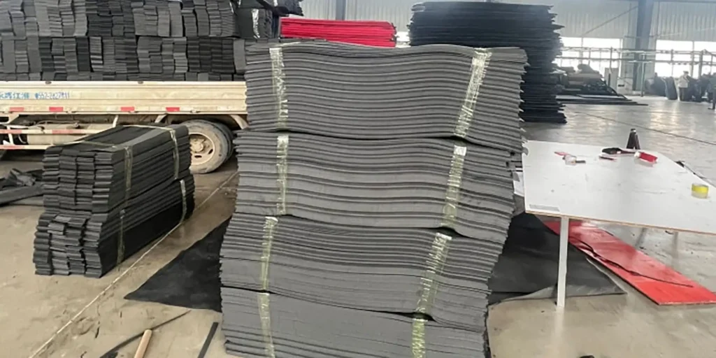 depicts a large-scale factory storage area where towering stacks of black industrial sheets (bound tightly with straps) dominate the scene. In the background, a truck (loaded with more materials), a worktable (with tools), and additional piles of black and red components are visible. The expansive, concrete-floored space and organized stacks indicate an active manufacturing or distribution environment for heavy-duty industrial materials.