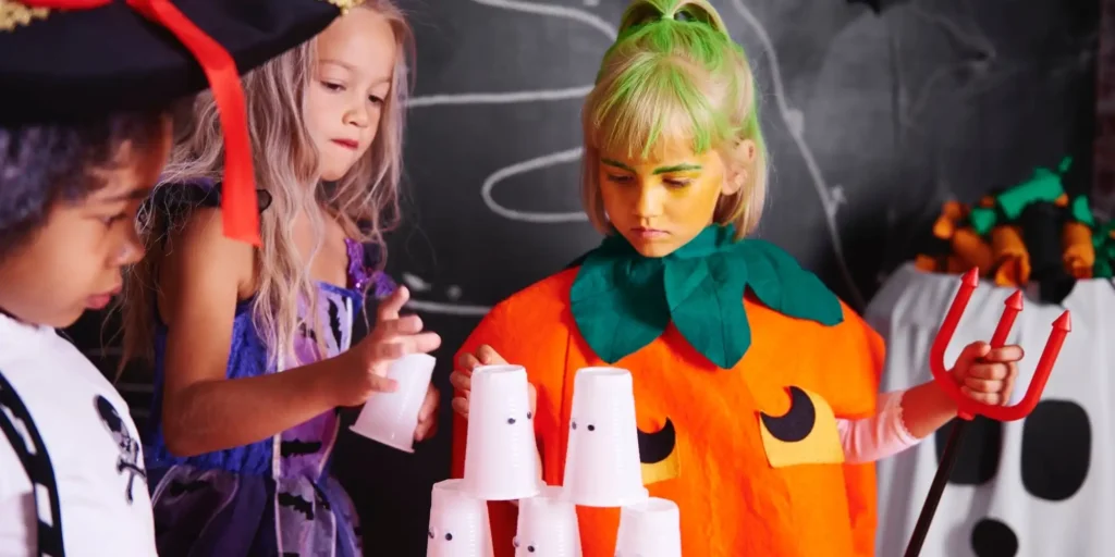 Three children in an indoor setting. The child on the left is wearing a pirate hat and a purple costume and is looking ahead intently. The child in the center has blonde hair and is wearing a witch-like costume with a green headband tied around her head and green and black makeup applied to her face. She appears to be playing a cupcake tower game and is holding a red plastic straw. The child on the right, on the other hand, is wearing an orange pumpkin costume with a black eye pattern and appears to be observing or preparing to join the game. The background is dark with some decorations such as large black and white polka dots and other holiday decorations.