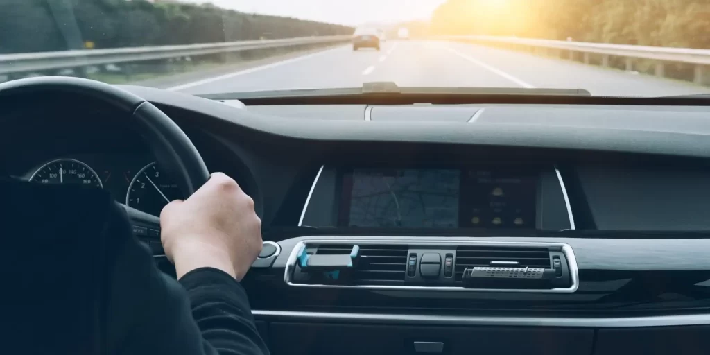 A man in a black jacket, driving a BMW X5, sits at the wheel, elbows resting on his knees, gazing out the window. The sun shines through the leaves as the vehicle travels down the road. His hair is wet with sweat and the collar of his shirt is a little damp.