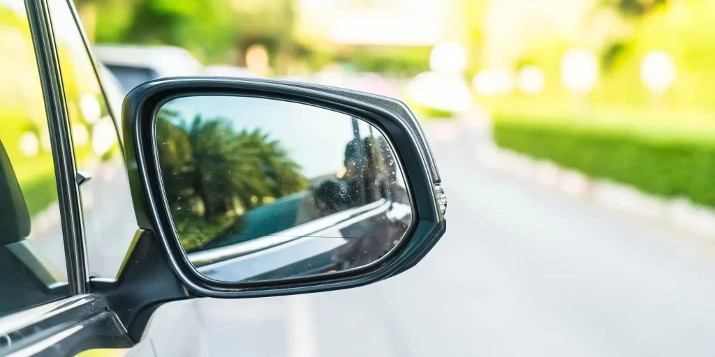 A woman is driving in the side view mirror of the car. She is wearing a black dress and white shawl, her hair tied casually in a ponytail. She sits in the driver's seat with her elbows on the steering wheel and her eyes on the street in front of her. Vehicles pass behind her and she seems to be focused on her driving process.