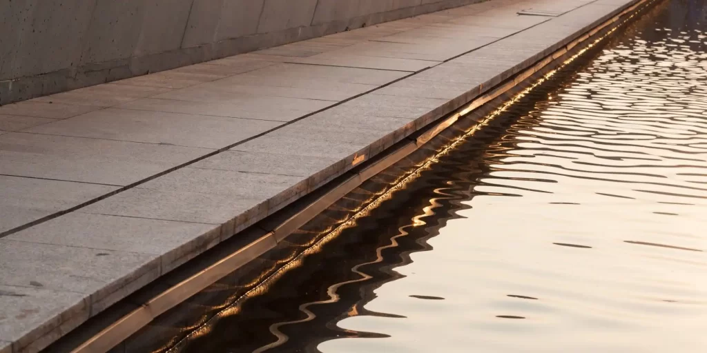 This picture shows a long gray stone path near the water's edge, next to calm water.