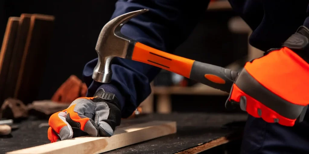 The picture shows a worker hammering a nail with an orange hammer, which penetrates a board. The worker is wearing black and orange gloves and the orange hammer has the Scott family logo on it.