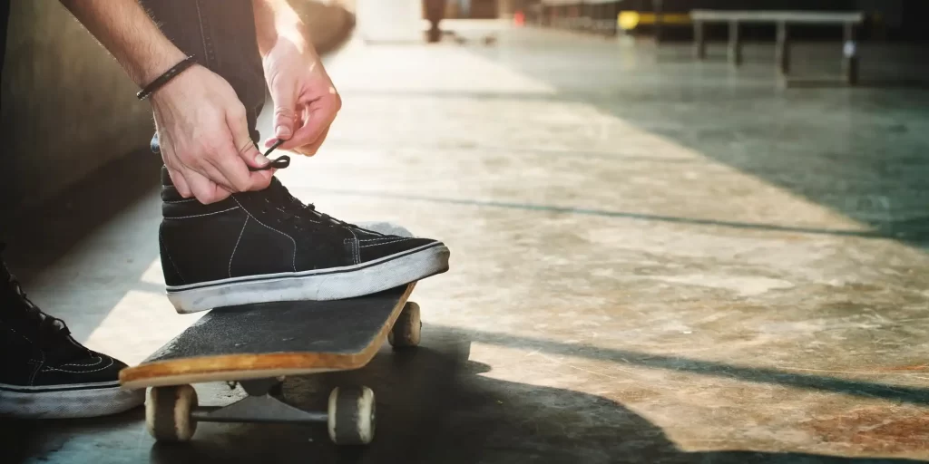 The image you've uploaded captures a close-up of someone tying the laces of black skateboarding shoes. The individual is standing with one foot on a skateboard, indicating that they are likely about to engage in skateboarding. Skateboarding shoes are typically designed with flat soles for better grip on the board, and the durability to withstand the wear and tear of skateboarding tricks and maneuvers. The setting appears to be an outdoor urban area, possibly a skate park or a street spot favored by skateboarders.
