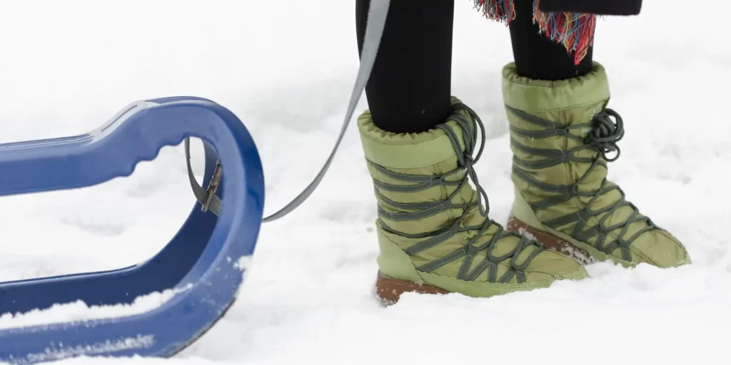 a close-up of someone's feet wearing green insulated winter boots, standing in the snow. There's also part of a blue sled in the foreground, suggesting that the setting might be a snowy hill suitable for sledding. These types of boots are designed to keep the feet warm and dry in snowy and icy conditions, often featuring thick insulation and waterproof materials. The boots' design suggests they are made for casual winter activities rather than for professional skiing, which usually requires specialized ski boots.