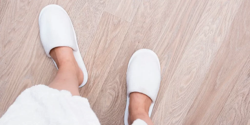 a pair of white slippers on someone's feet, standing on a wooden floor. The slippers appear soft and comfortable, typically associated with indoor wear. They look like they are made of a material that could include EVA foam, which is known for its softness and durability, providing comfort for the wearer.