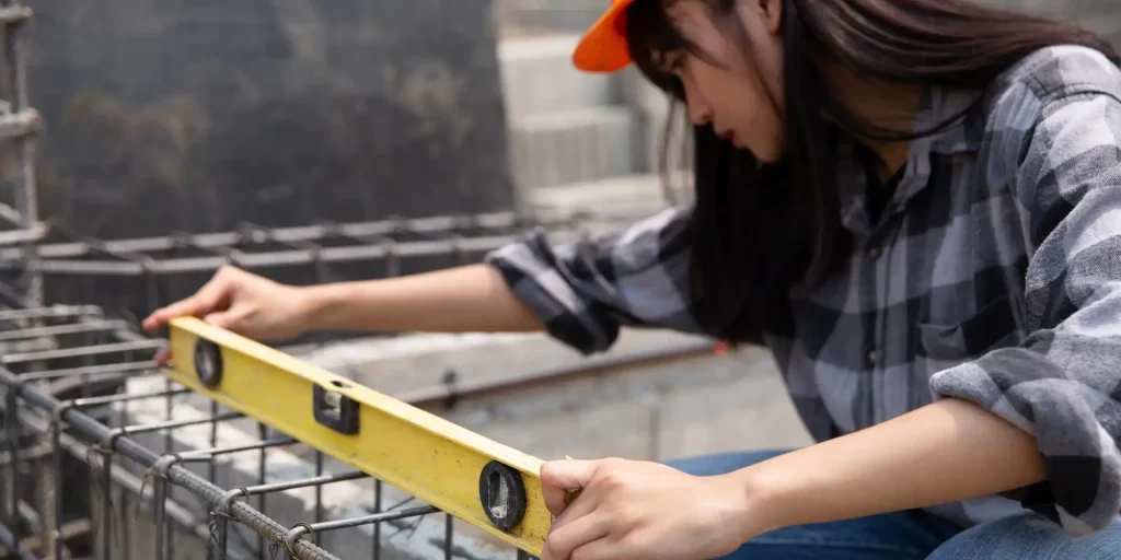 The picture shows a female construction worker, wearing an orange hat and plaid shirt, measuring a metal frame using a level.