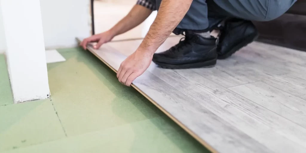 The picture shows a worker installing flooring, kneeling on the floor with a piece of flooring in his hand. The floor is light brown in color with a smooth and wood grain textured surface. The floor in the worker's hand has a gap between it and the wall for filling and fixing.