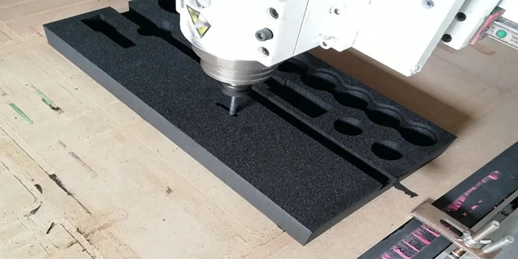 AlternativeText: A CNC machine precisely cutting a black foam material with pre - existing cutouts. Caption: CNC Machine in Action: Precision Cutting of Black Foam Description: This image showcases a CNC machine actively engaged in cutting a black foam material. The foam features multiple pre - formed cutouts, highlighting a meticulous, precision - oriented process. The machine, characterized by its white body and metallic components, is positioned above the foam, exemplifying industrial - level manufacturing. The surrounding workspace includes a cardboard - like surface, indicating a controlled environment designed for accurate cutting operations.