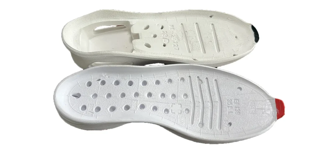 two white shoe soles placed one above the other. Both soles are designed with numerous holes and grooves, which likely serve purposes such as breathability and flexibility. The upper sole has a black toe tip, while the lower one has a red toe tip. Additionally, there are engraved markings on each sole, including alphanumeric codes, which may indicate specifications or manufacturing details. The overall appearance suggests they are components intended for footwear construction.