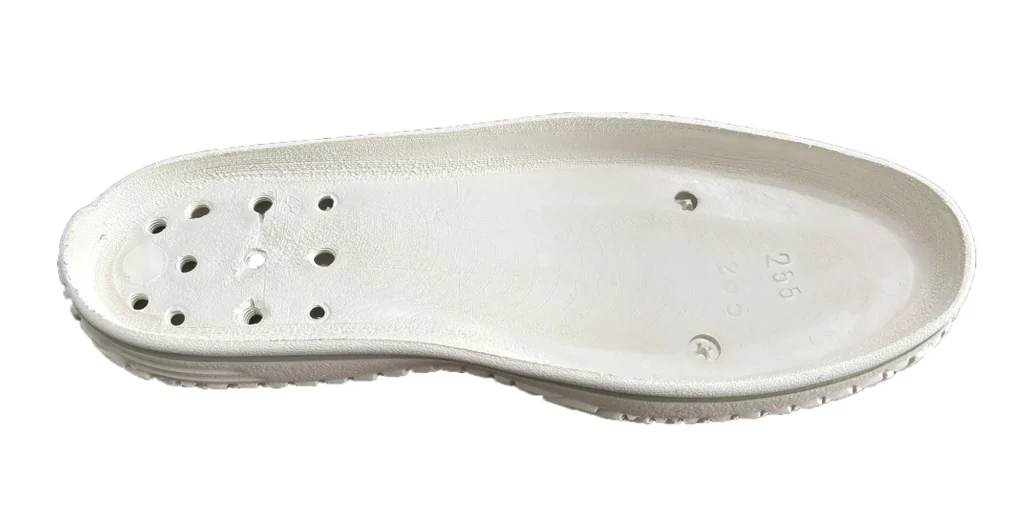 a sole has a textured surface and is designed with multiple holes, which may contribute to breathability. There are also some engraved markings on the sole, including numbers and symbols, likely indicating specifications or part of the manufacturing details. The overall appearance suggests it is a component intended for footwear construction.