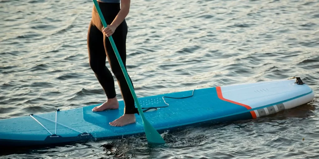 Woman standing on surfboard with paddle in hand. She is wearing a wetsuit and lifejacket and standing in the waves. She appears to be paddling or swimming, surrounded by the sound of the ocean's waves. She swims freely through the water, weaving over the waves like a fish. Her body posture is graceful and confident, showing her freedom and vigor.