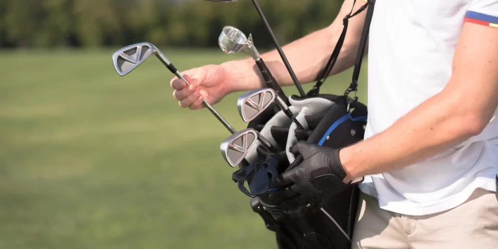 The man is holding golf clubs and wearing a white shirt and black pants, surrounded by green grass and a blue sky with white clouds. He confidently displays his professionalism and love for the game of golf, holding several golf clubs in his hands as he appears to be practicing his swinging skills. The man shows the charm of the sport and a positive spirit.