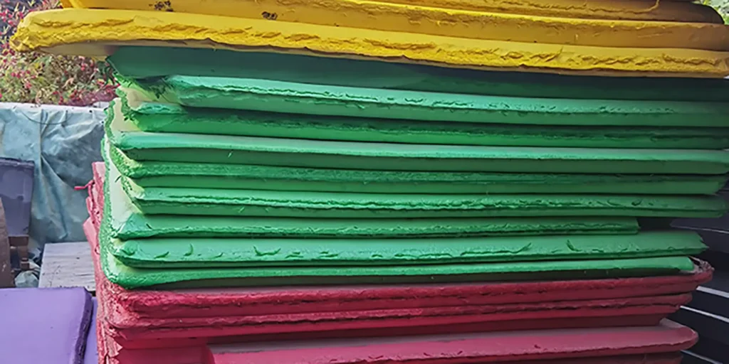 depicts an outdoor storage scene where a tall stack of industrial sheets is arranged in distinct color layers: yellow at the top, followed by green, and red at the bottom. The sheets have a slightly textured surface, and the background includes covered materials (likely other industrial components), indicating a functional storage environment for these colorful, sturdy materials (possibly for manufacturing or craft applications).